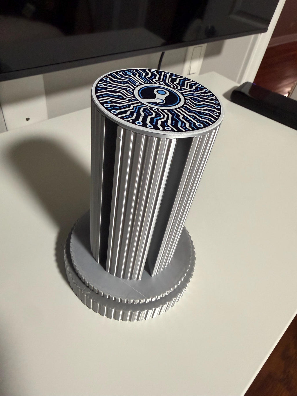 Steam Machine Faceplate Organizer, Spinning 3D Printed Display Stand, Rotating Plate Holder, Gamer Desk Setup, Gift for Gamer, Holds 6, top-down angled view of the empty silk silver organizer column standing alone on a white desk with a dark monitor behind, showing the circuit-board patterned top cap with the Steam logo