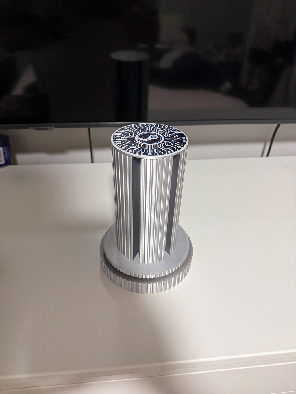 Steam Machine Faceplate Organizer, Spinning 3D Printed Display Stand, Rotating Plate Holder, Gamer Desk Setup, Gift for Gamer, Holds 6, empty silk silver organizer column standing alone centered on a white desk seen from the front, showing the circuit-board patterned top cap with the Steam logo, with a dark TV behind