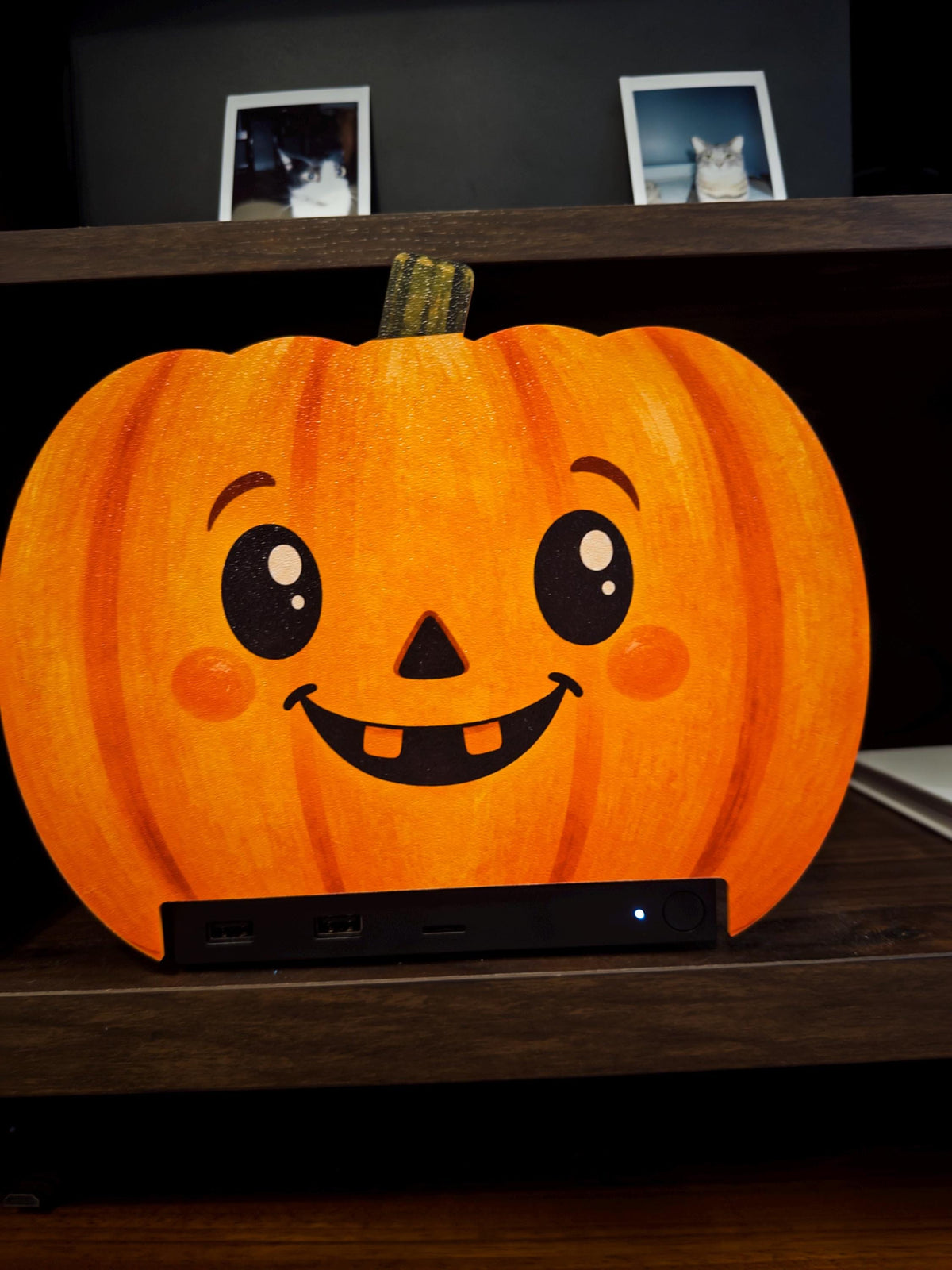 Steam Machine Faceplate Halloween Pumpkin, Classic or Cute Jack o Lantern Magnetic Plate, UV Printed Console Cover, Spooky Gamer Gif, close-up front view of the cute round pumpkin faceplate with big shiny eyes, rosy cheeks and two teeth, installed on the Steam Machine on a dark wooden shelf, with two framed cat photos on the shelf above