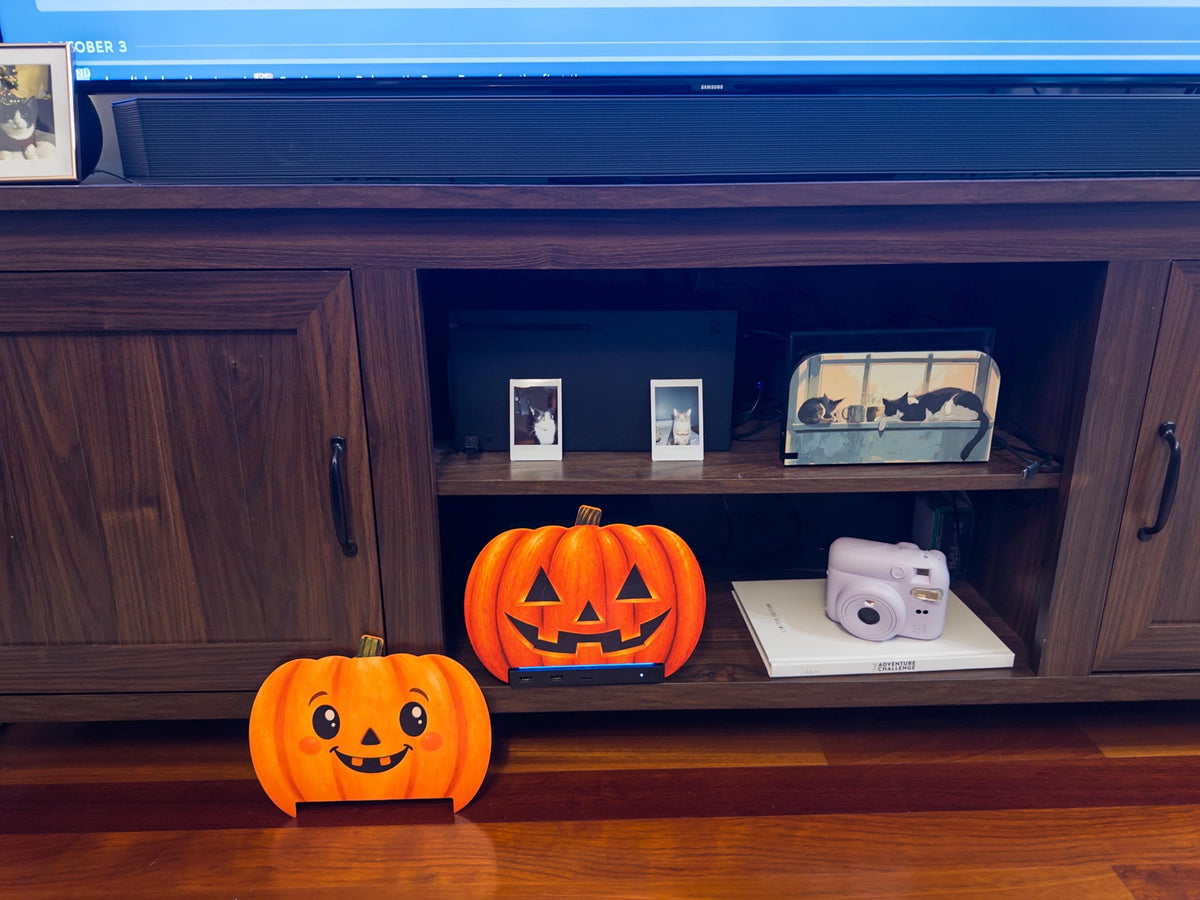 Steam Machine Faceplate Halloween Pumpkin, Classic or Cute Jack o Lantern Magnetic Plate, UV Printed Console Cover, Spooky Gamer Gif, both pumpkin faceplate designs side by side, with the classic carved jack-o-lantern faceplate installed on the Steam Machine on a TV stand shelf and the cute pumpkin faceplate standing on the wooden floor in front, beside a lavender instant camera on a book