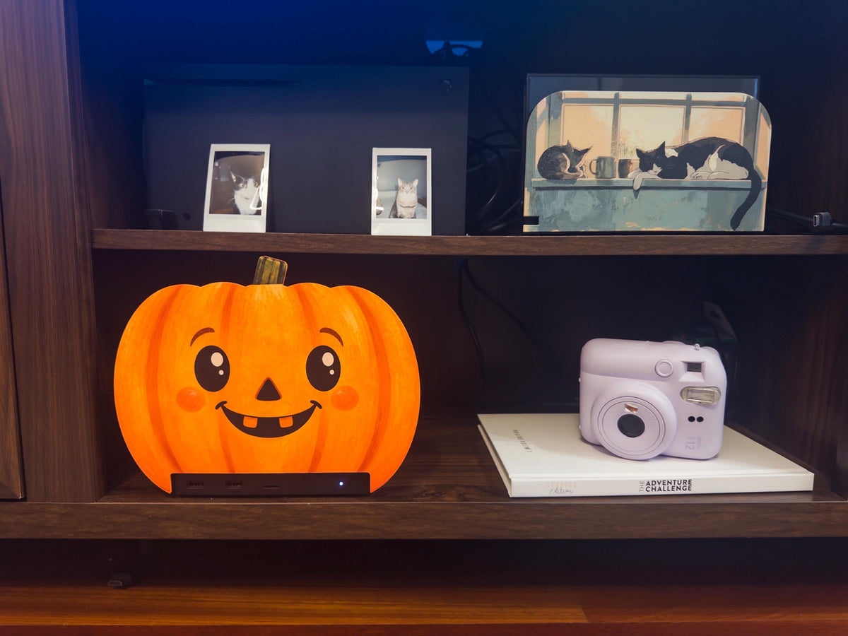 Steam Machine Faceplate Halloween Pumpkin, Classic or Cute Jack o Lantern Magnetic Plate, UV Printed Console Cover, Spooky Gamer Gif, cute round pumpkin faceplate installed on the Steam Machine on a dark wooden shelf, beside a lavender instant camera on a white book, with framed cat photos and a window-cats artwork on the shelf above
