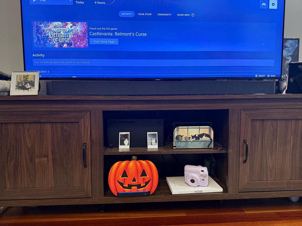 Steam Machine Faceplate Halloween Pumpkin, Classic or Cute Jack o Lantern Magnetic Plate, UV Printed Console Cover, Spooky Gamer Gif, classic carved jack-o-lantern pumpkin faceplate installed on the Steam Machine on the lower shelf of a dark wood media console, beneath a TV showing a game library screen, with a lavender instant camera on a book beside it