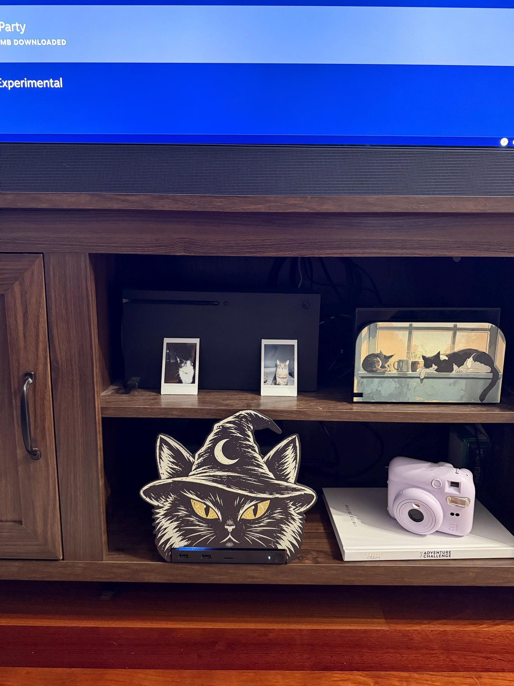 Steam Machine Faceplate Black Cat Witch Hat, Halloween Witchy Cat Magnetic Plate, UV Printed Console Cover, Spooky Gamer Gift, black cat witch hat faceplate installed on the Steam Machine on a wooden media cabinet shelf beside a pink instant camera, with a TV above and small framed cat photos on the shelf above