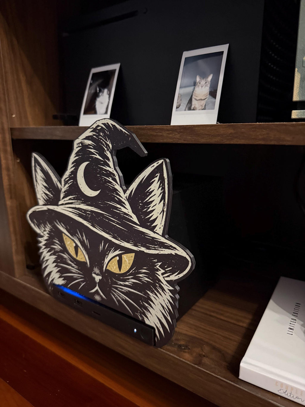 Steam Machine Faceplate Black Cat Witch Hat, Halloween Witchy Cat Magnetic Plate, UV Printed Console Cover, Spooky Gamer Gift, angled view of the black cat witch hat faceplate installed on the Steam Machine on a wooden shelf, with a white book beside it and two instant cat photo prints on the shelf above