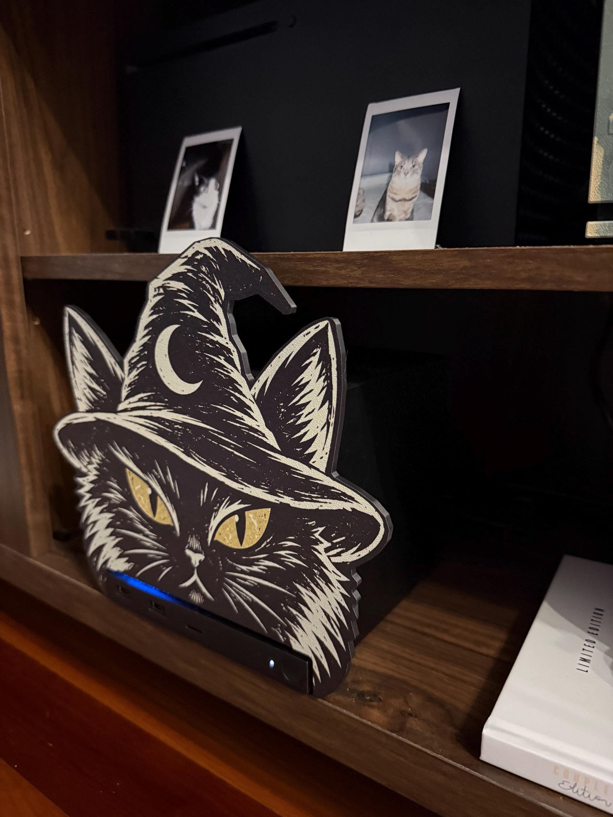 Steam Machine Faceplate Black Cat Witch Hat, Halloween Witchy Cat Magnetic Plate, UV Printed Console Cover, Spooky Gamer Gift, angled view of the black cat witch hat faceplate installed on the Steam Machine on a wooden shelf, with a white book beside it and two instant cat photo prints on the shelf above