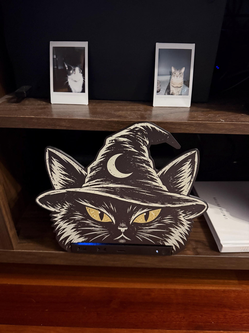 Steam Machine Faceplate Black Cat Witch Hat, Halloween Witchy Cat Magnetic Plate, UV Printed Console Cover, Spooky Gamer Gift, front view of the black cat witch hat faceplate installed on the Steam Machine on a wooden shelf, with two instant cat photo prints on the dark wall behind and a book beside it