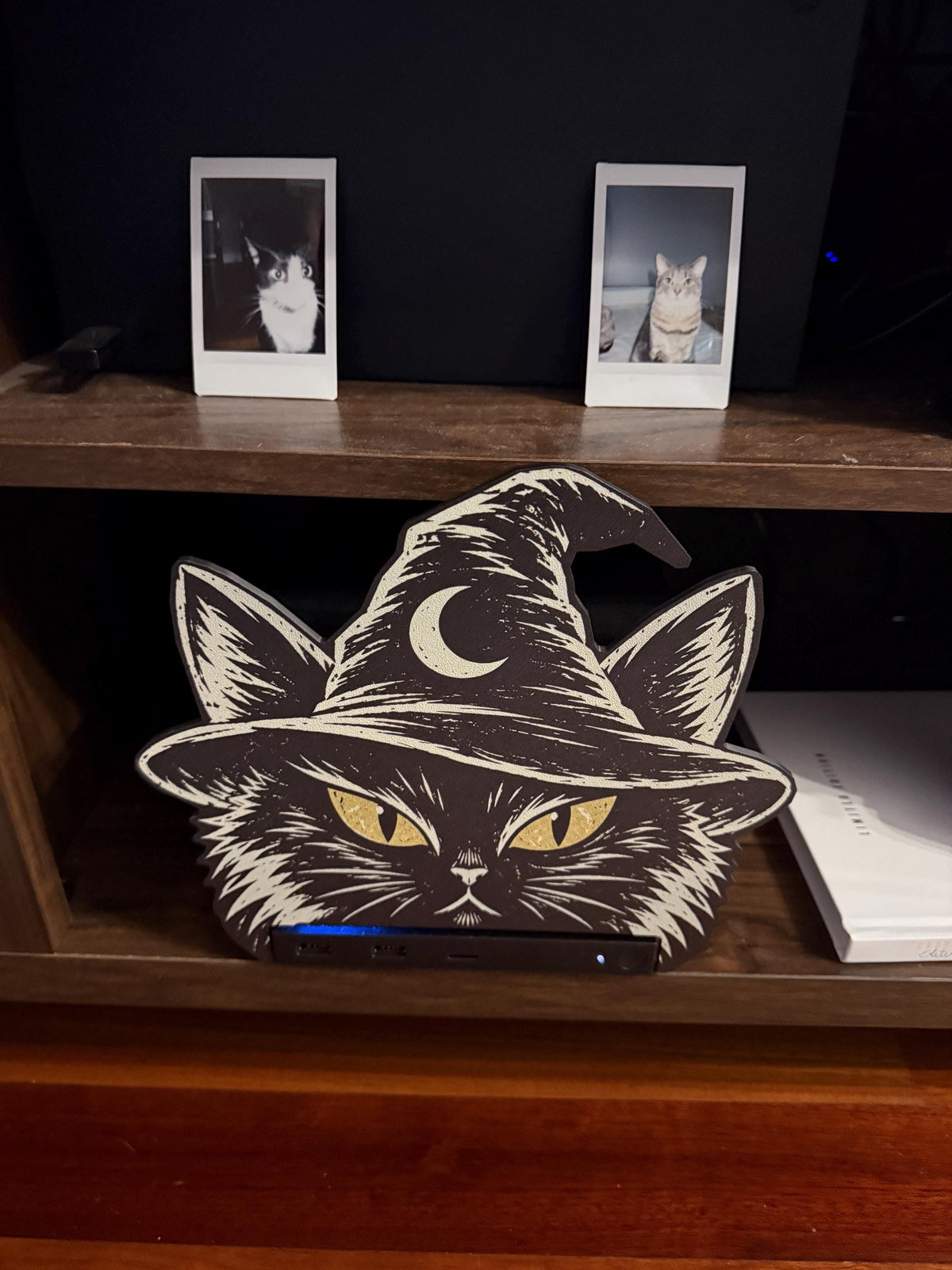 Steam Machine Faceplate Black Cat Witch Hat, Halloween Witchy Cat Magnetic Plate, UV Printed Console Cover, Spooky Gamer Gift, front view of the black cat witch hat faceplate installed on the Steam Machine on a wooden shelf, with two instant cat photo prints on the dark wall behind and a book beside it
