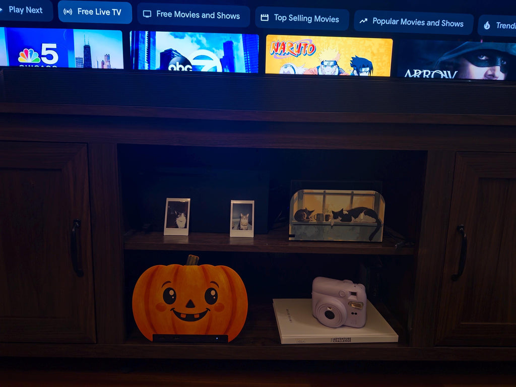Steam Machine Faceplate Halloween Pumpkin, Classic or Cute Jack o Lantern Magnetic Plate, UV Printed Console Cover, Spooky Gamer Gif, wide view of a dark wood media console with a TV above showing streaming tabs, and the cute pumpkin faceplate installed on the Steam Machine on the lower open shelf beside a lavender instant camera on a book