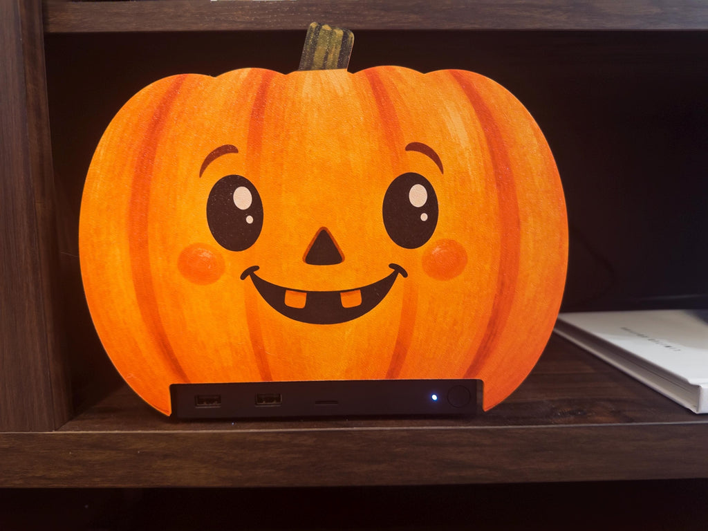 Steam Machine Faceplate Halloween Pumpkin, Classic or Cute Jack o Lantern Magnetic Plate, UV Printed Console Cover, Spooky Gamer Gif, close-up view of the cute round pumpkin faceplate with big shiny eyes, rosy cheeks and two teeth, installed on the Steam Machine on a wooden shelf beside a white book