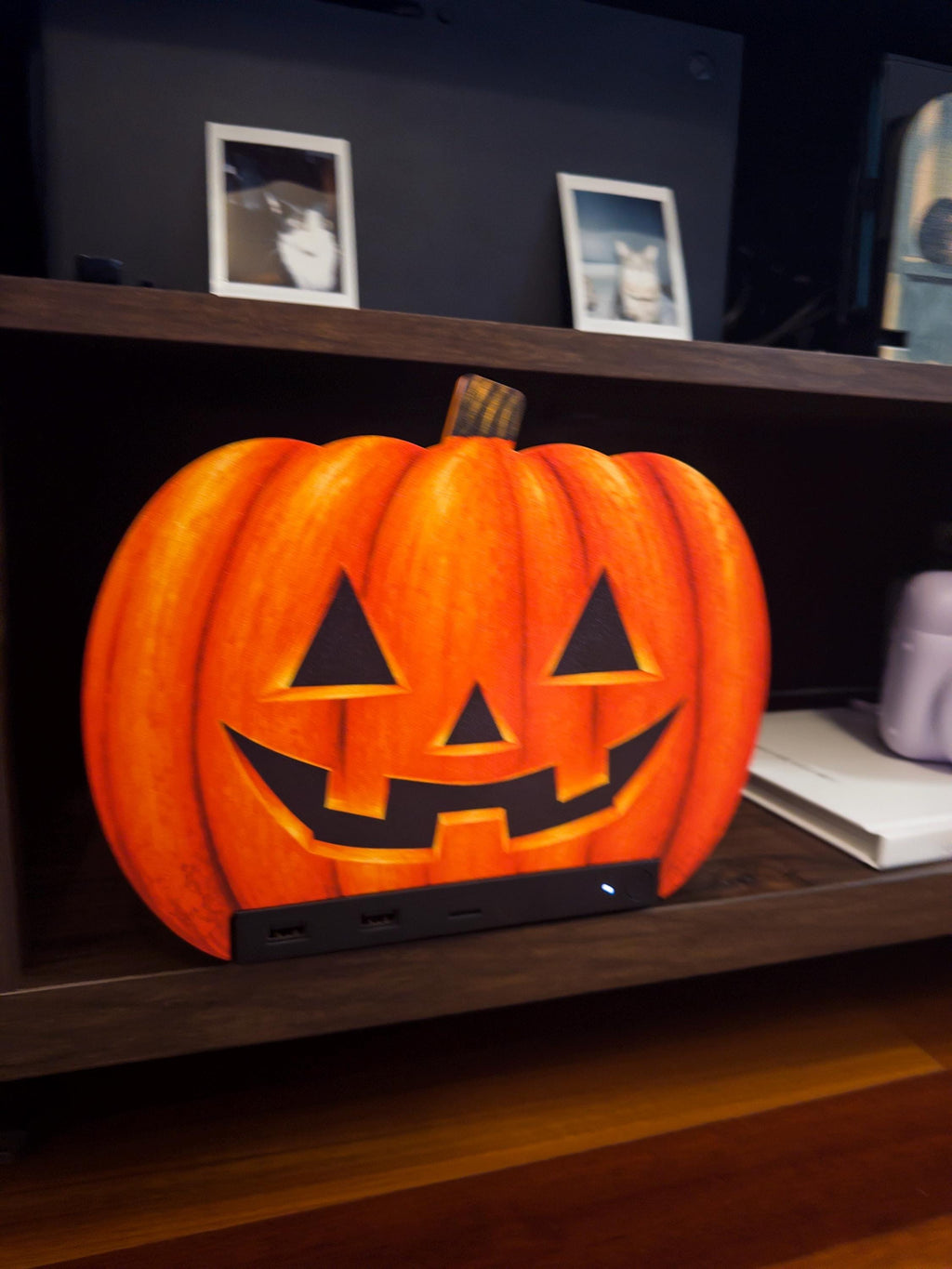 Steam Machine Faceplate Halloween Pumpkin, Classic or Cute Jack o Lantern Magnetic Plate, UV Printed Console Cover, Spooky Gamer Gif, angled close-up view of the classic carved jack-o-lantern pumpkin faceplate installed on the Steam Machine on a wooden shelf, with a lavender instant camera on a white book beside it and framed cat photos on the shelf above