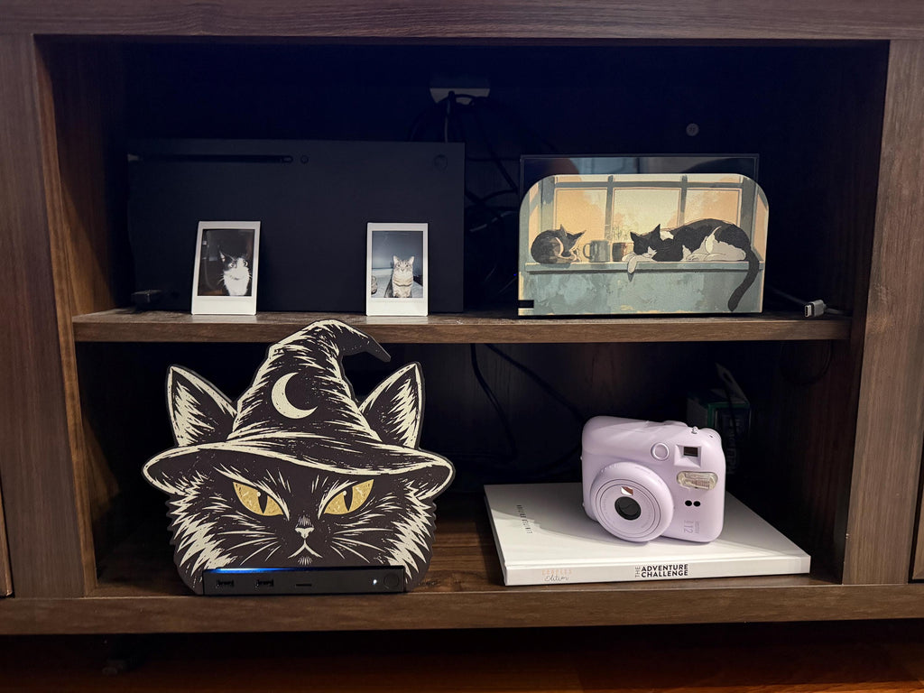 Steam Machine Faceplate Black Cat Witch Hat, Halloween Witchy Cat Magnetic Plate, UV Printed Console Cover, Spooky Gamer Gift, wooden cabinet shelf with the black cat witch hat faceplate installed on the Steam Machine beside a lavender instant camera, with two instant cat photo prints and a framed window cat picture on the shelf above