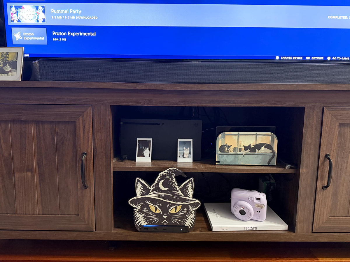 Steam Machine Faceplate Black Cat Witch Hat, Halloween Witchy Cat Magnetic Plate, UV Printed Console Cover, Spooky Gamer Gift, wide view of a dark brown wooden media console with the black cat witch hat faceplate installed on the Steam Machine in the center open shelf, beside a lavender instant camera, with a large TV above showing a blue interface