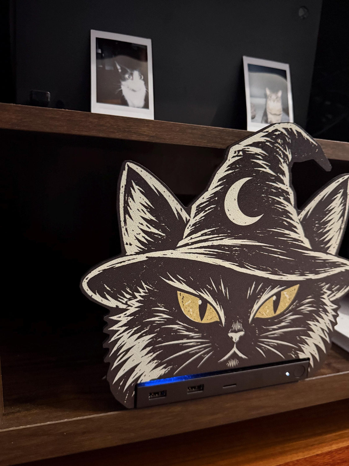 Steam Machine Faceplate Black Cat Witch Hat, Halloween Witchy Cat Magnetic Plate, UV Printed Console Cover, Spooky Gamer Gift, close-up of the black cat witch hat faceplate installed on the Steam Machine on a wooden shelf, with framed black-and-white cat photos hanging on the dark wall behind