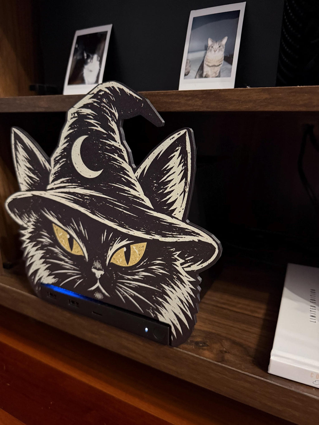 Steam Machine Faceplate Black Cat Witch Hat, Halloween Witchy Cat Magnetic Plate, UV Printed Console Cover, Spooky Gamer Gift, angled close-up of the black cat witch hat faceplate installed on the Steam Machine on a wooden shelf, with two instant cat photo prints on the dark wall behind and a book on the shelf below