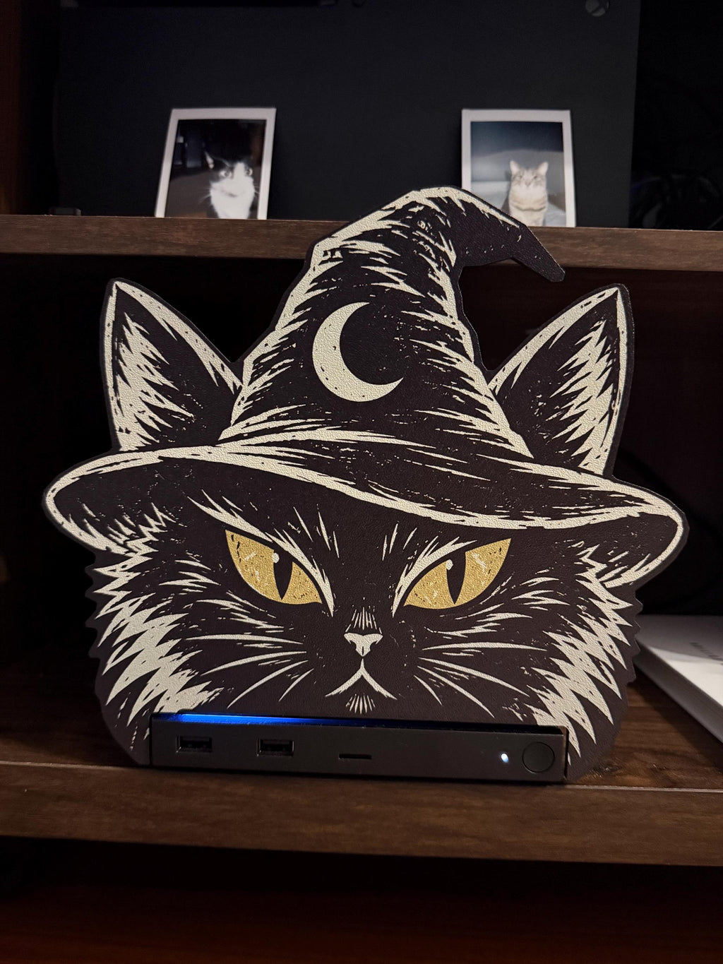 Steam Machine Faceplate Black Cat Witch Hat, Halloween Witchy Cat Magnetic Plate, UV Printed Console Cover, Spooky Gamer Gift, front view of the black cat witch hat faceplate installed on the Steam Machine on a wooden shelf, showing the console's blue light strip and USB ports below, with two instant cat photo prints on the dark wall behind