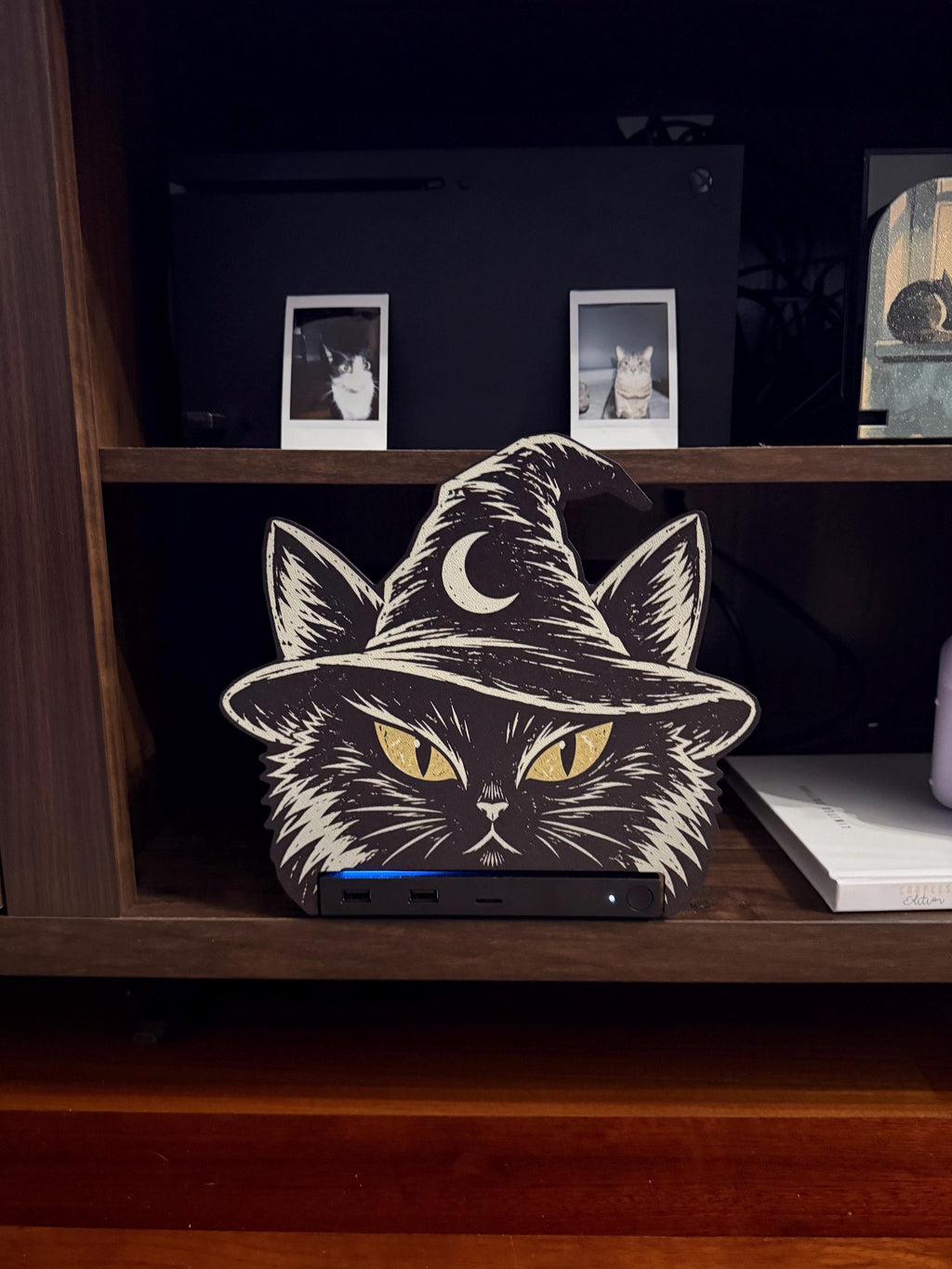 Steam Machine Faceplate Black Cat Witch Hat, Halloween Witchy Cat Magnetic Plate, UV Printed Console Cover, Spooky Gamer Gift, front view of the black cat witch hat faceplate installed on the Steam Machine on a wooden shelf, with a white book beside it and two instant cat photo prints on the shelf above