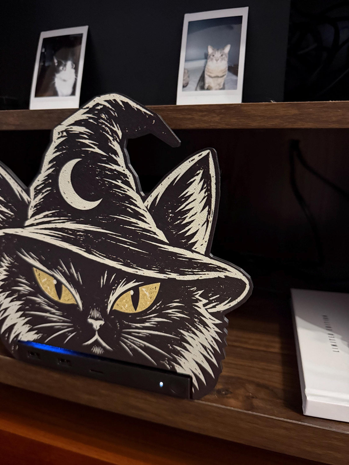 Steam Machine Faceplate Black Cat Witch Hat, Halloween Witchy Cat Magnetic Plate, UV Printed Console Cover, Spooky Gamer Gift, angled view from the left of the black cat witch hat faceplate installed on the Steam Machine, showing the console's blue light strip, with a white book beside it and two instant cat photo prints on the shelf above