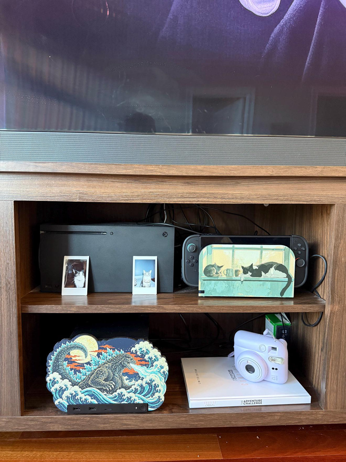 Steam Machine Faceplate, Custom Faceplate, Godzilla Kaiju Plate XL Oversized, Magnetic Console Cover, Monster Gamer Gift, Japan Wave Decor, portrait view of the Godzilla kaiju XL faceplate installed on the open shelf of a wooden TV console below a television and soundbar, beside a white book with a lavender instant camera